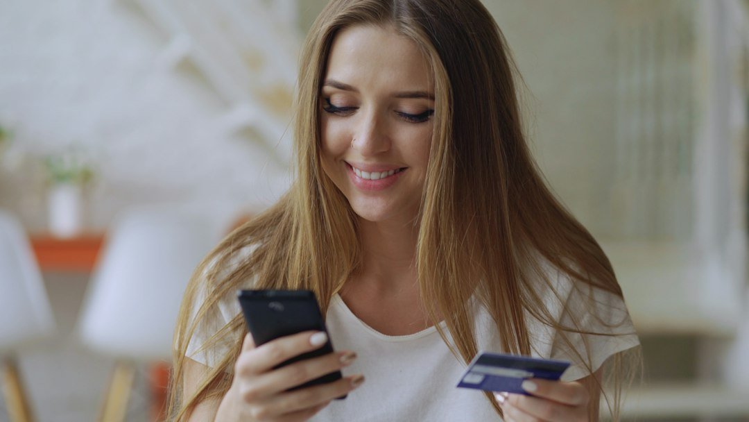 A woman shopping online using a smartphone and debit card at home, representing agentic AI shopping conflicts
