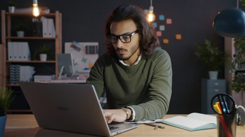 Man working on a laptop and taking notes at night, doing a remote side hustle after his day job