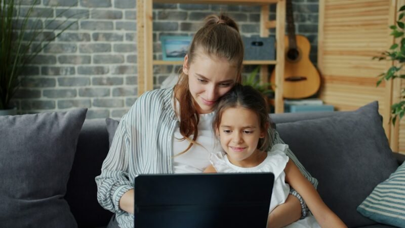 Mother using a laptop at home with her young daughter, a side hustle for stay-at-home moms