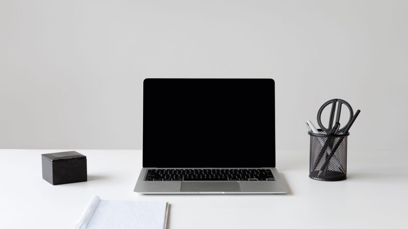 A laptop on a white desk next to a notepad and pen holder