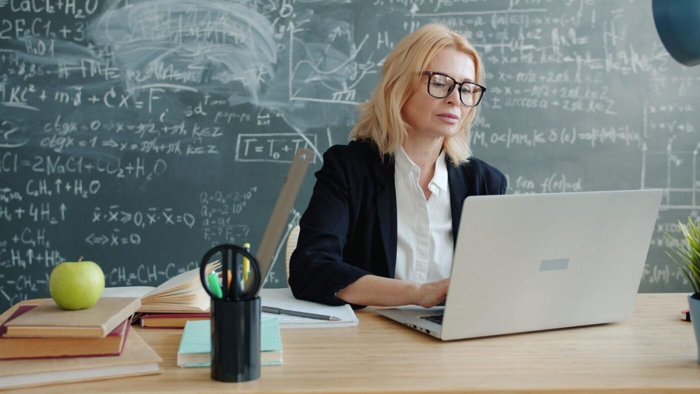 Teacher working on a laptop at a desk in front of a chalkboard