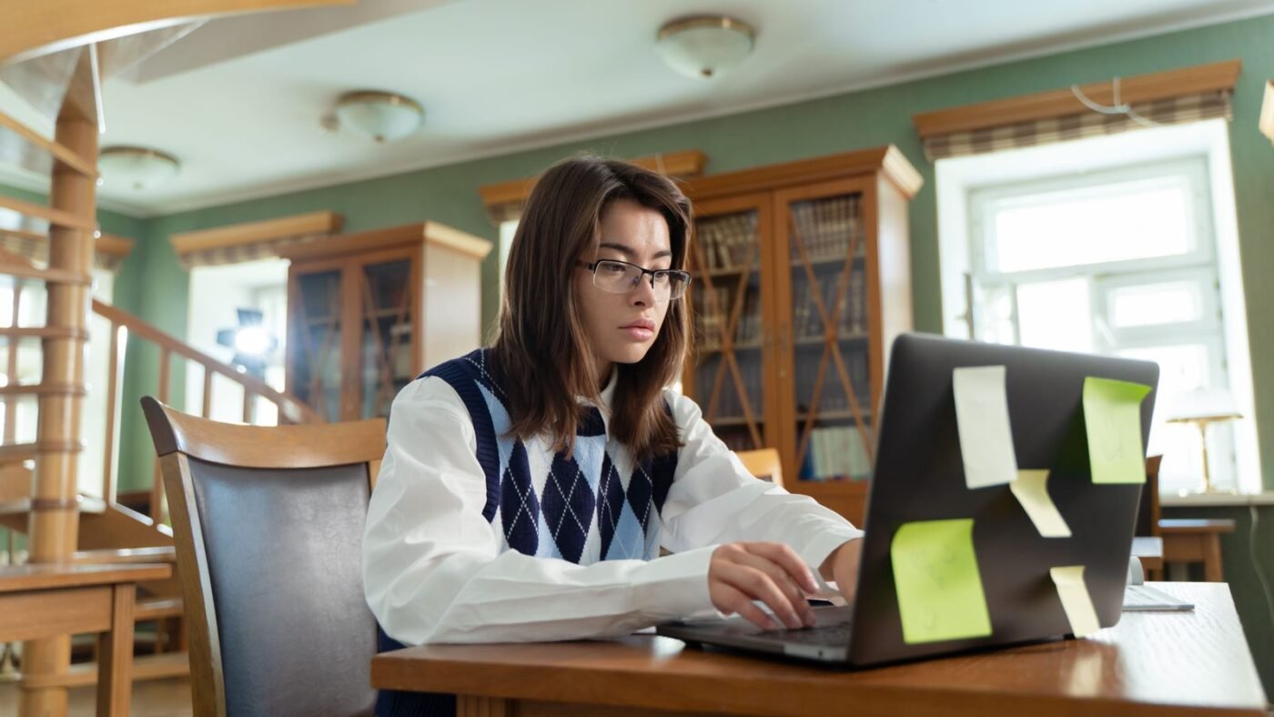 Student typing on a laptop at a wooden desk in a library