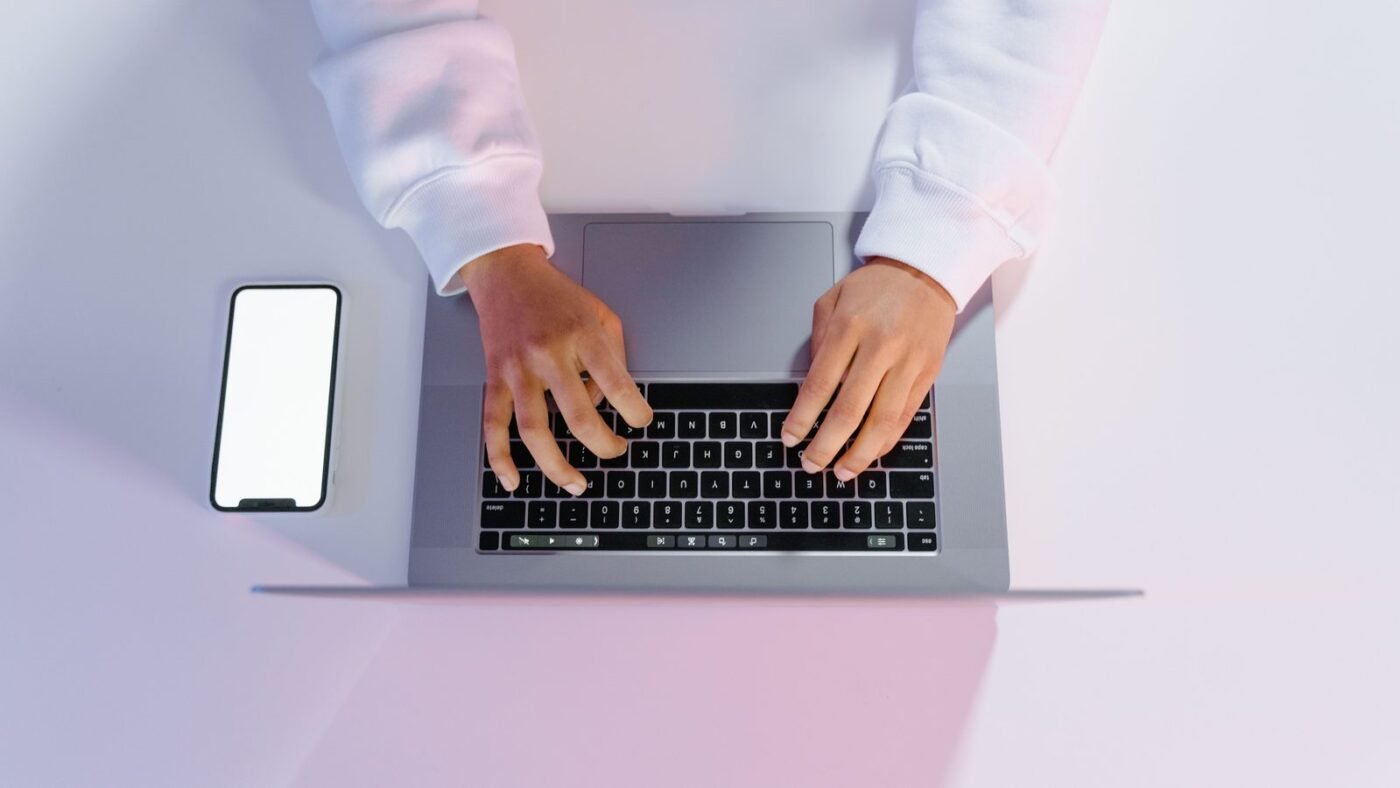 Hands typing on a laptop beside a smartphone on a minimalist white desk
