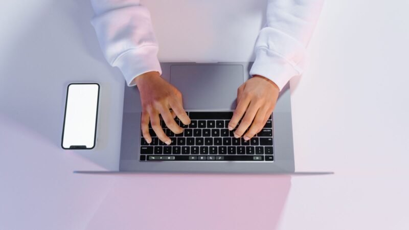 Hands typing on a laptop beside a smartphone on a minimalist white desk