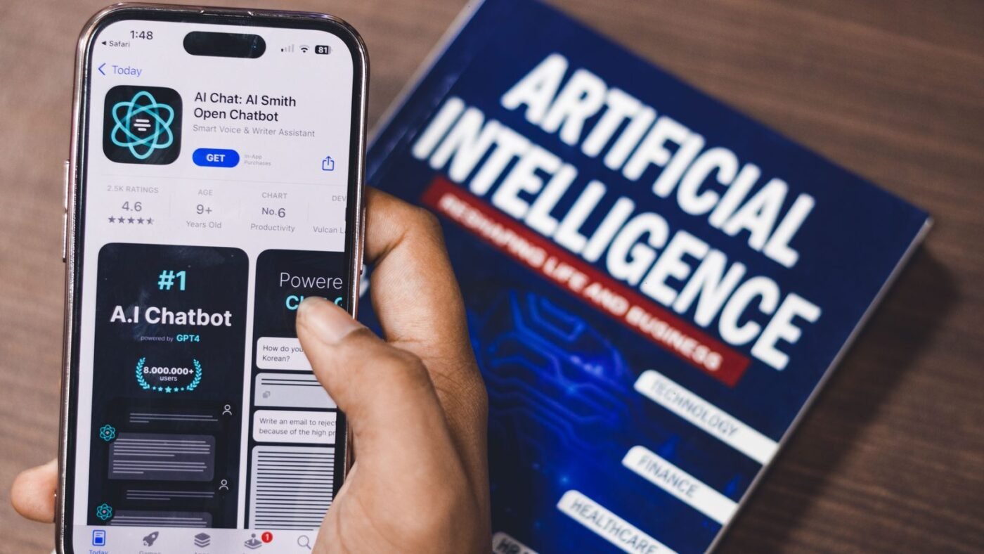 Hand holding a smartphone showing an AI chatbot app beside a book about artificial intelligence