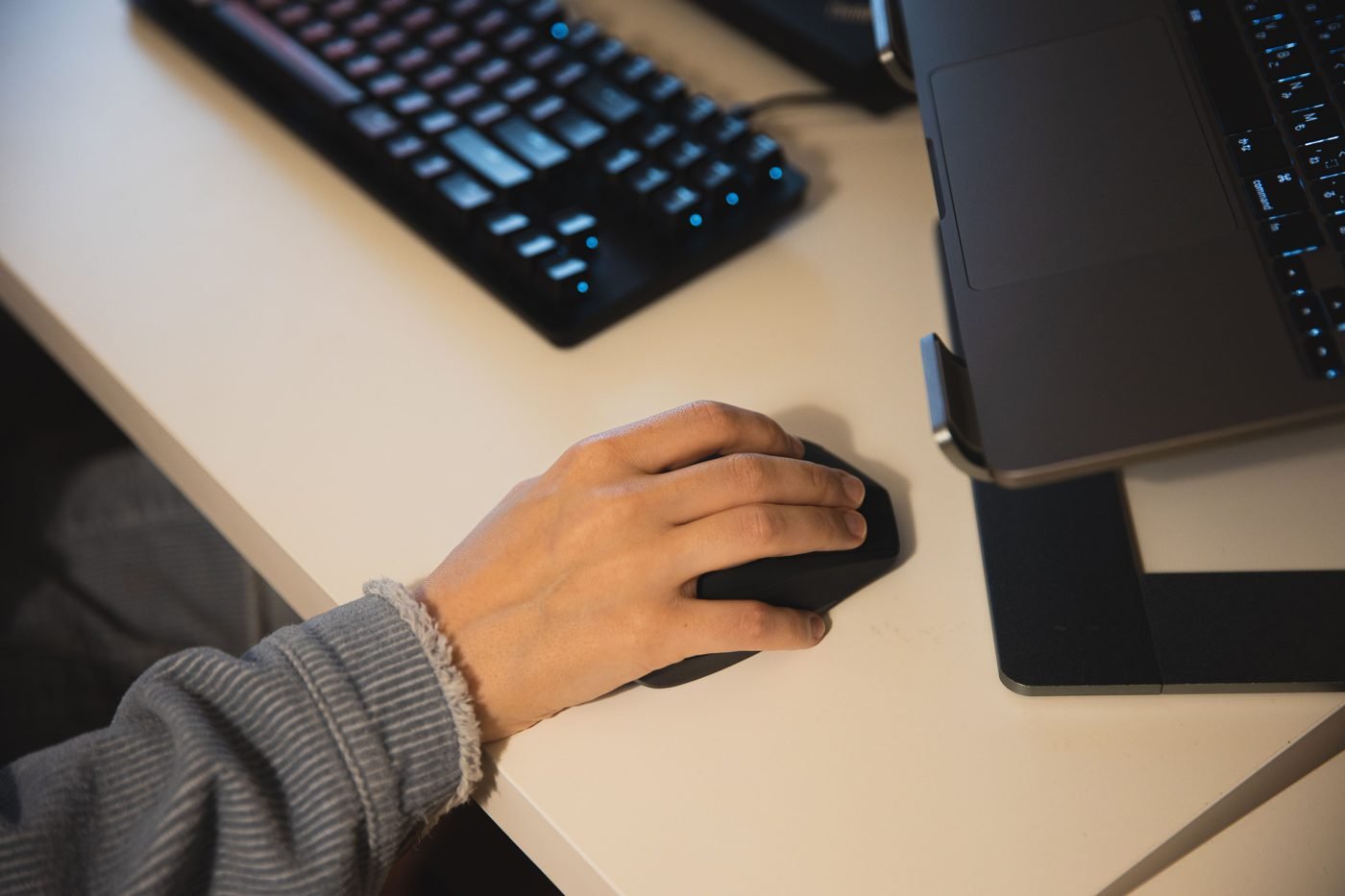Hand using a computer mouse next to a laptop and keyboard on a desk