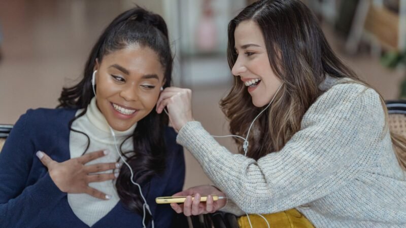 Two friends smiling and sharing earphones while listening to music on a smartphone