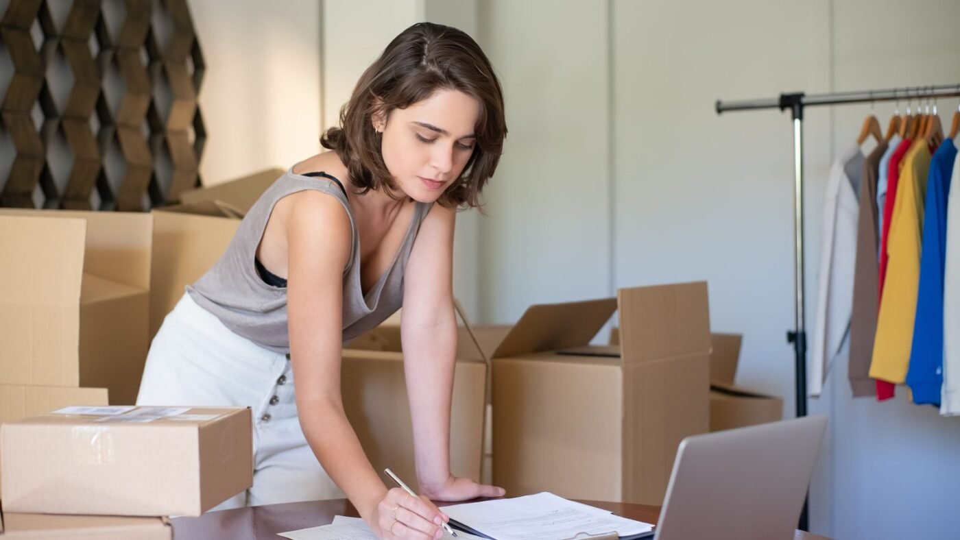 A small business owner writing notes at a desk surrounded by shipping boxes