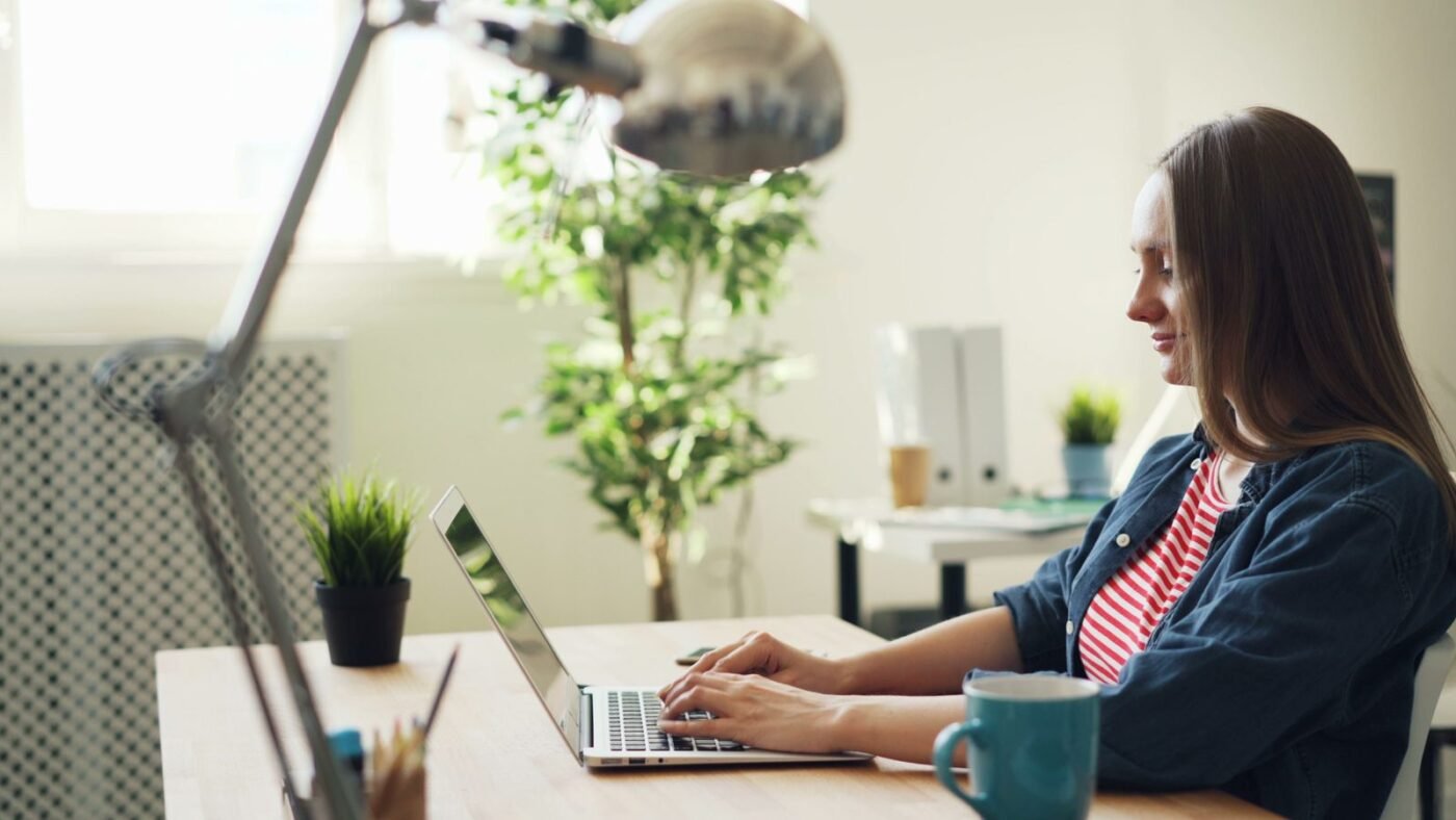 Woman smiling while working on a laptop at a bright office desk with plants