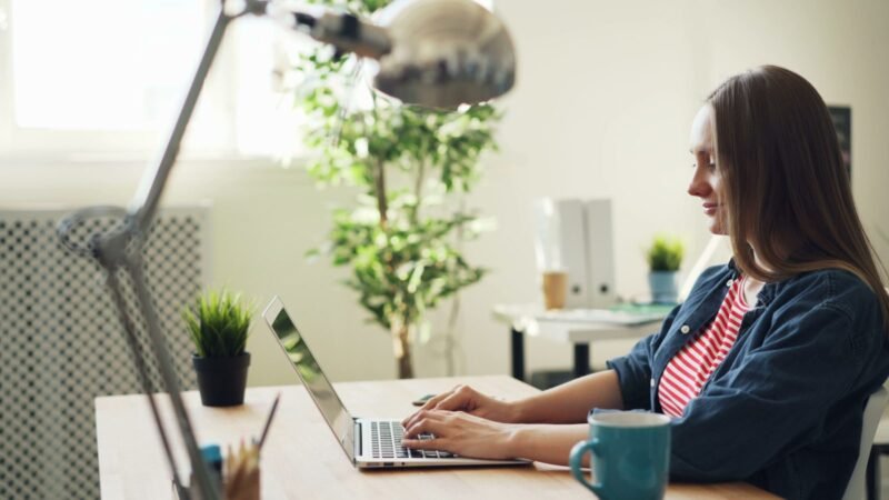 Woman smiling while working on a laptop at a bright office desk with plants