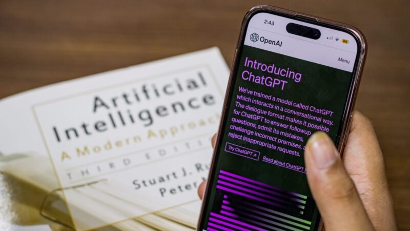 Hand holding a smartphone showing the ChatGPT app above an artificial intelligence textbook