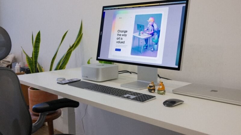 Desktop workstation with a large monitor, keyboard and a compact computer on a white desk beside indoor plants