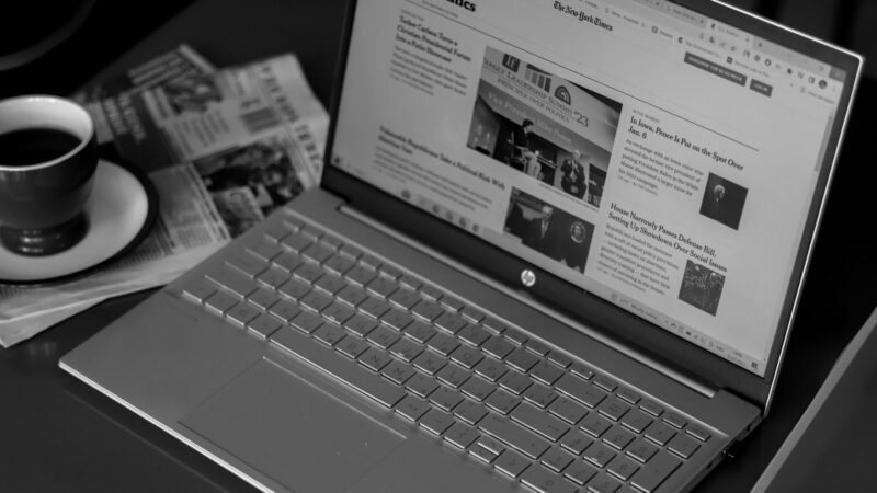 Black and white photo of a laptop displaying news articles beside a newspaper and coffee