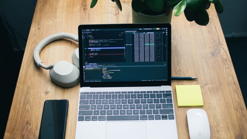 Laptop and headphones on a wooden desk next to a smartphone and a houseplant