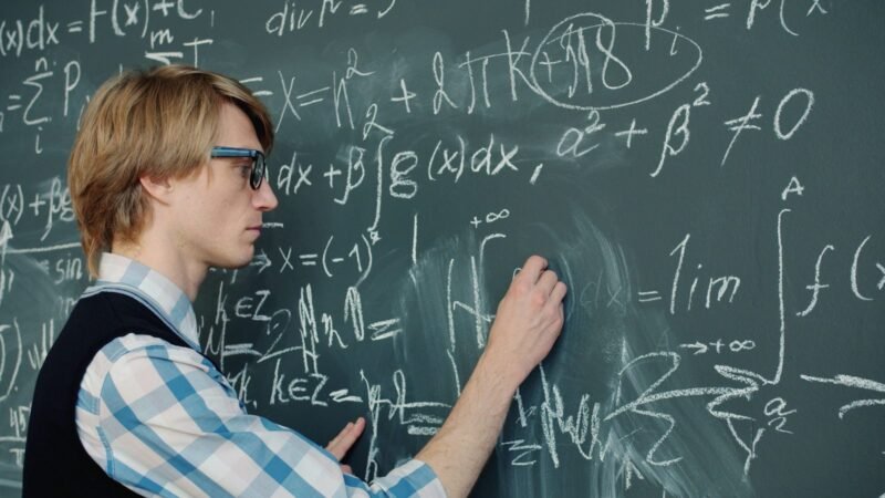 Young man in glasses writing equations on a green chalkboard