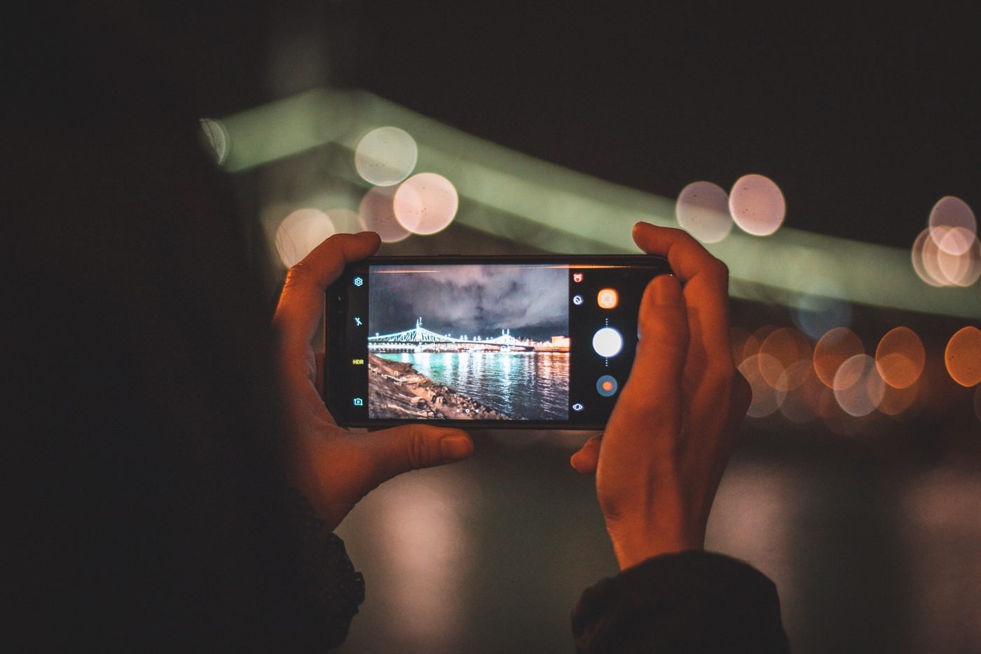 Hands holding a smartphone photographing a city skyline at night