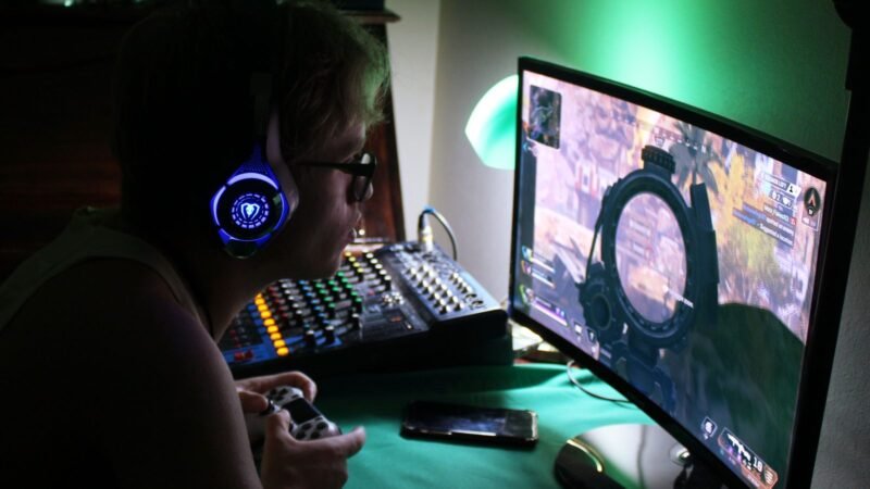 Gamer wearing a headset and holding a controller in a dark room lit by a monitor