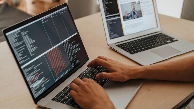 Developer typing code on a laptop next to a second laptop on a wooden desk