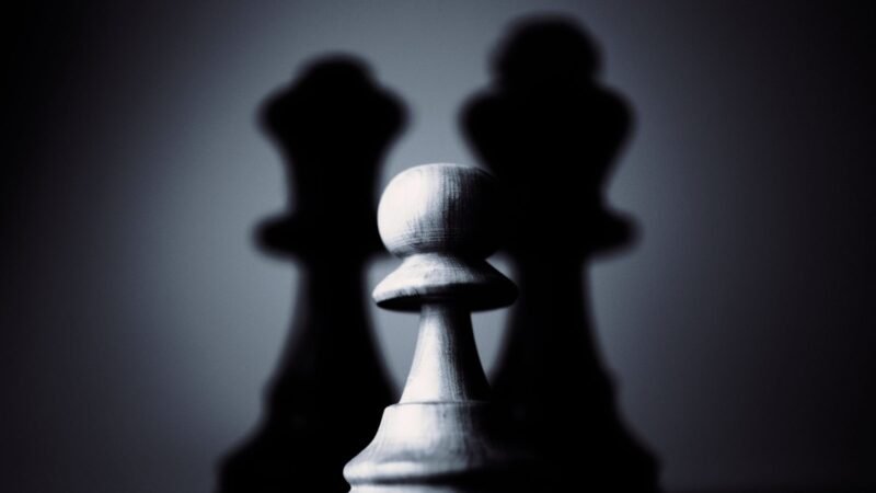 White chess pawn in front of dark chess pieces in dramatic lighting