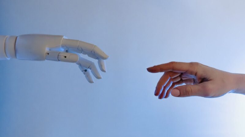A robotic hand reaching toward a human hand against a pale blue background