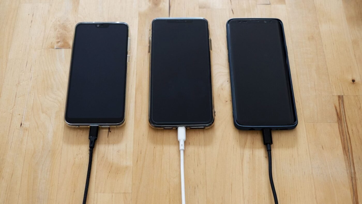 Three smartphones plugged into charging cables on a wooden table